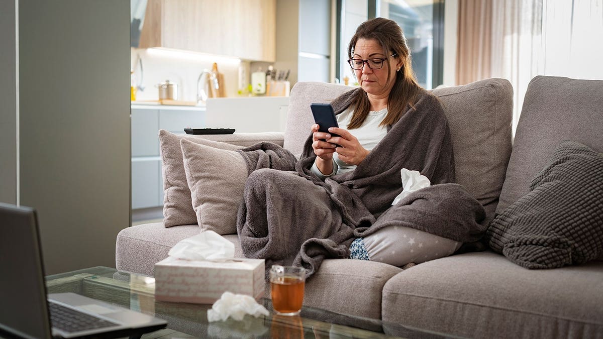Woman rests on sofa at home using smartphone, wrapped in blanket.
