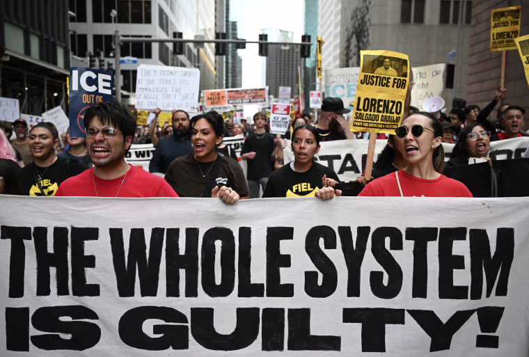 Protestors holding a large white banner reading THE WHOLE SYSTEM IS GUILTY!
