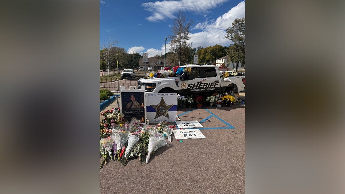 Memorial honoring Deputy Katherine Frank features a white sheriff's pickup truck covered with flowers, framed photographs, bouquets and handwritten tributes outside a law enforcement building.