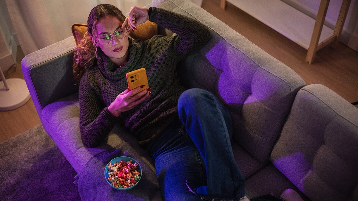 Young woman enjoying a relaxing evening at home, using her smartphone while lying on a comfortable sofa and eating popcorn