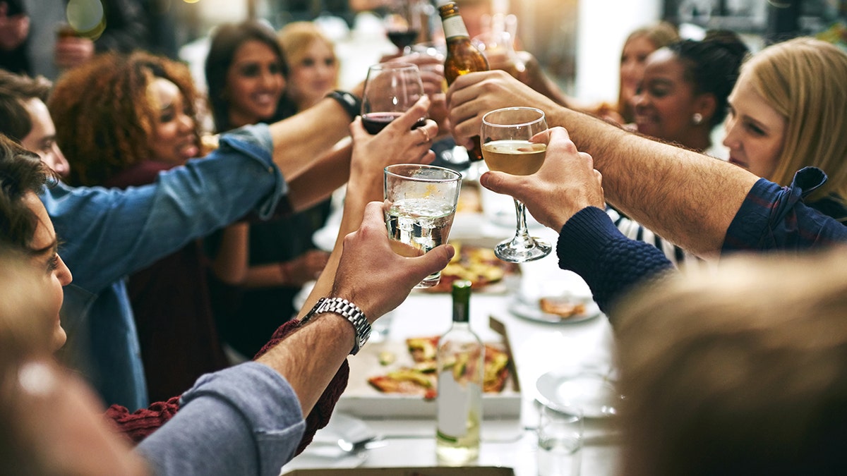 Group of young people toasting drinks together at long table in restaurant, food seen along table.