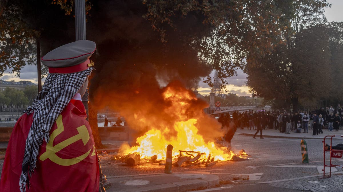 A protester wearing a hammer-and-sickle flag watches a fire burn at a demonstration in Paris, France, on Saturday, Oct. 17, 2026. Rioters attacked French public buildings and looted stores in cities including Paris as protests against the high cost of living drew more than half a million people, underscoring the nation's challenges ahead of presidential elections next year.