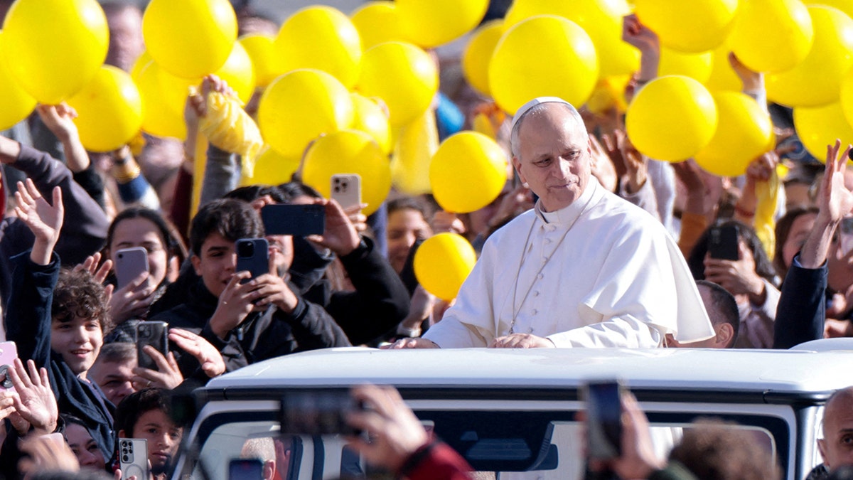 Pope Leo XIV waving to a crowd from a vehicle in a large square.