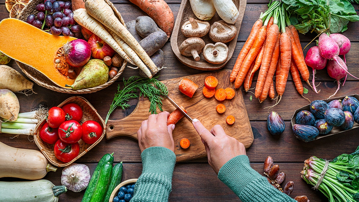 Woman chopping carrots on a table full of healthy Autumnal fruits and vegetables. Fresh organic seasonal produce background. Healthy vegetarian raw food. Food that boost the body inmune system. Nutrition and wellness, healthy diet and vegetable choice. Overhead view