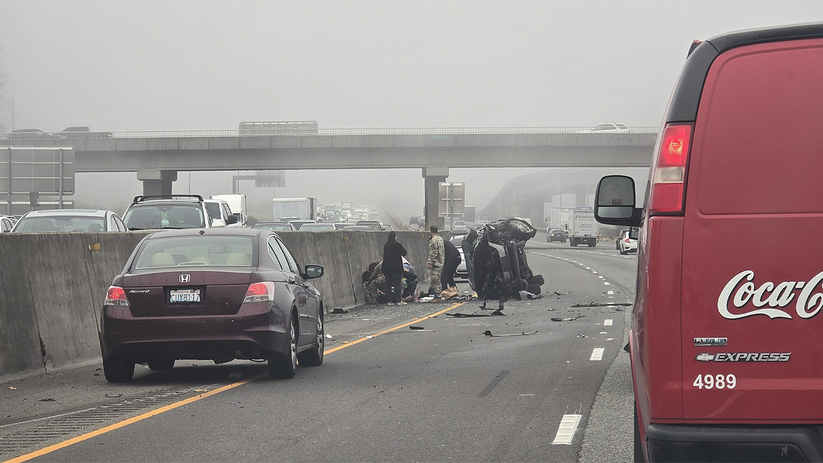 People in military uniforms and other bystanders assist beside an overturned SUV with debris scattered across I-5