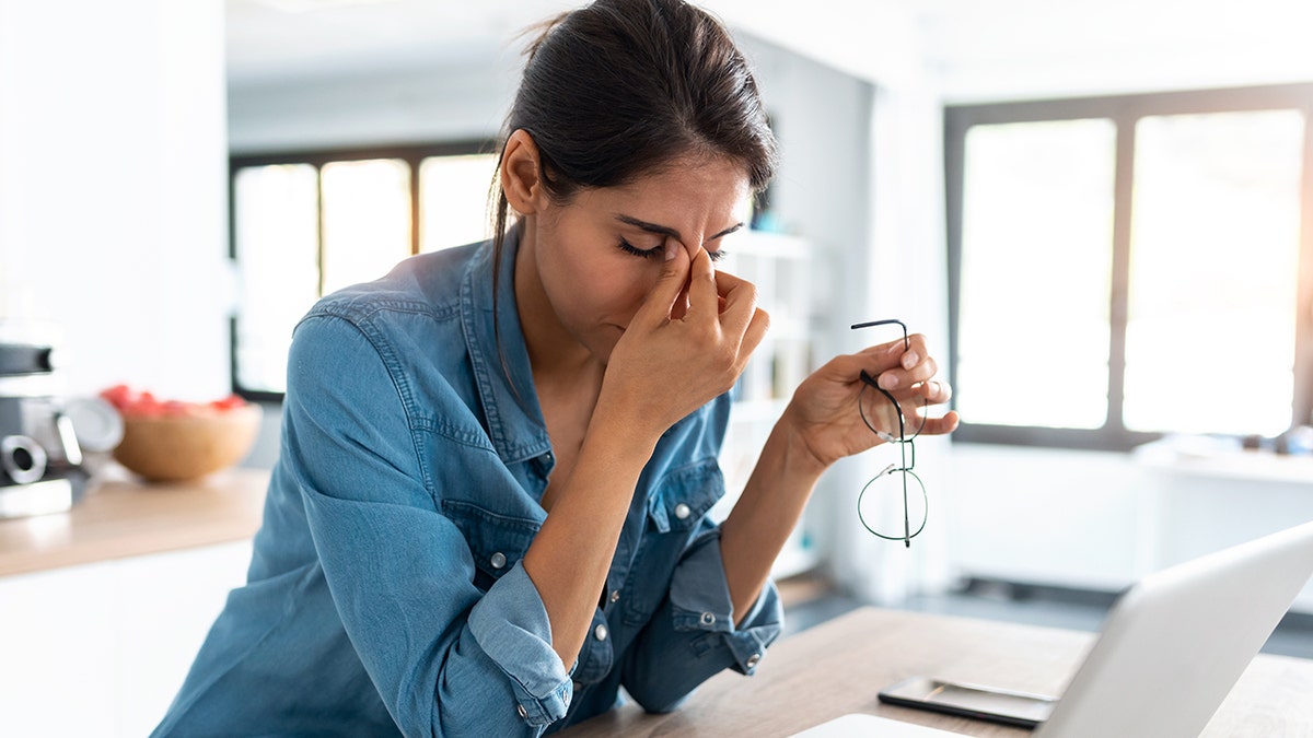 Woman in denim shirt pinching her nose in stress while holding glasses in front of a laptop at home.